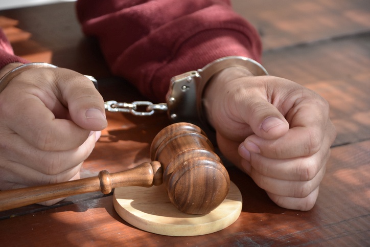 A gavel sits on a wooden surface alongside a pair of hands cuffed in steel restraints. The image evokes the legal themes of justice, guilt, and potential abuse of power.